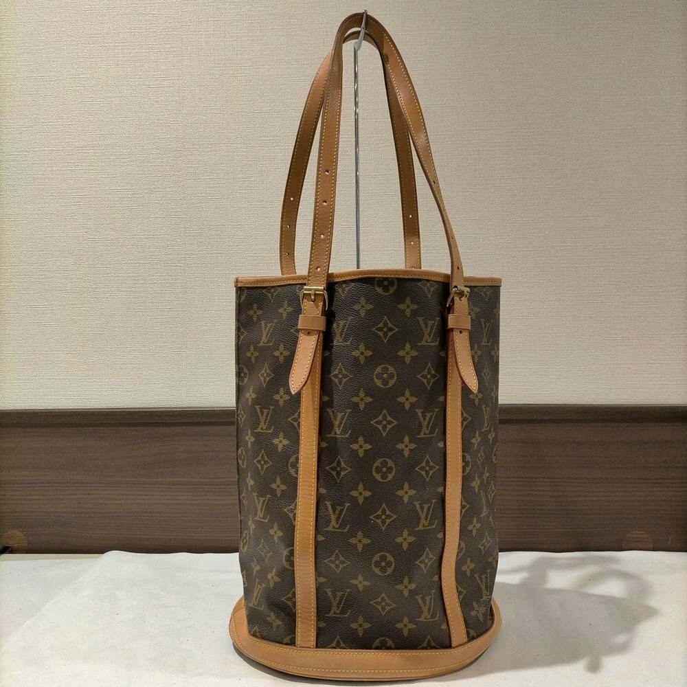 Front view of a brown Louis Vuitton monogram bucket bag with leather trim and double shoulder straps standing upright.