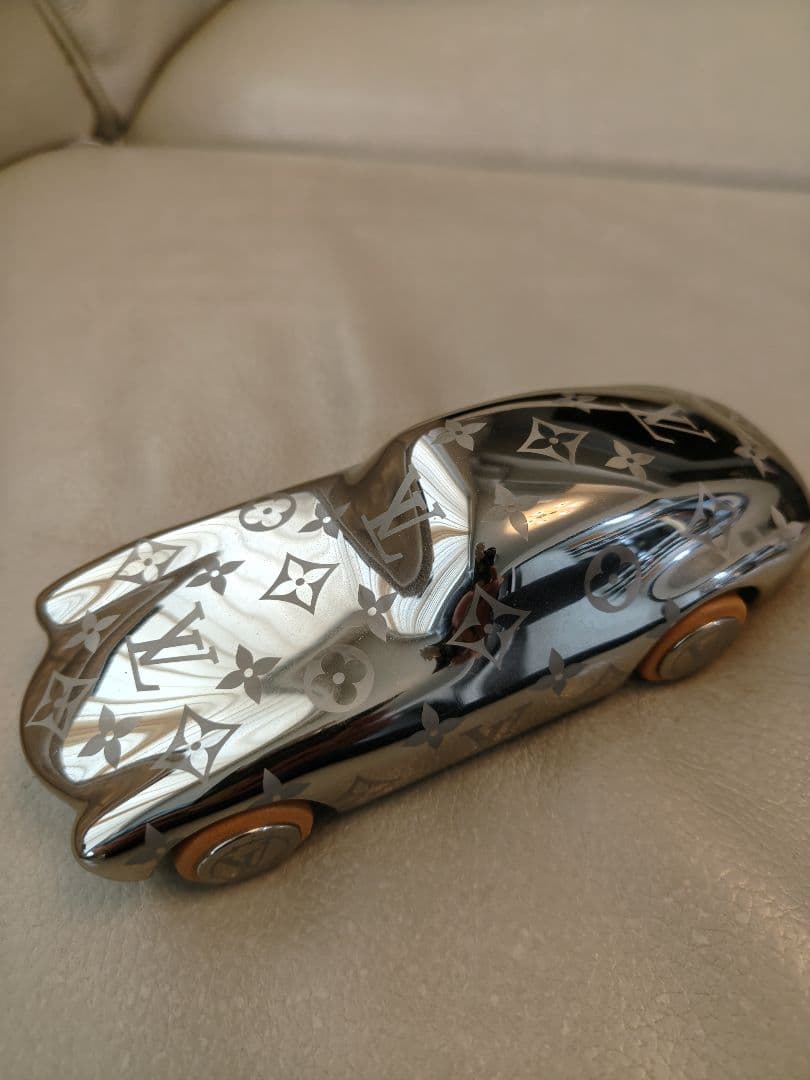 A reflective Louis Vuitton monogram car-shaped brass paperweight with visible wheels on a light surface.