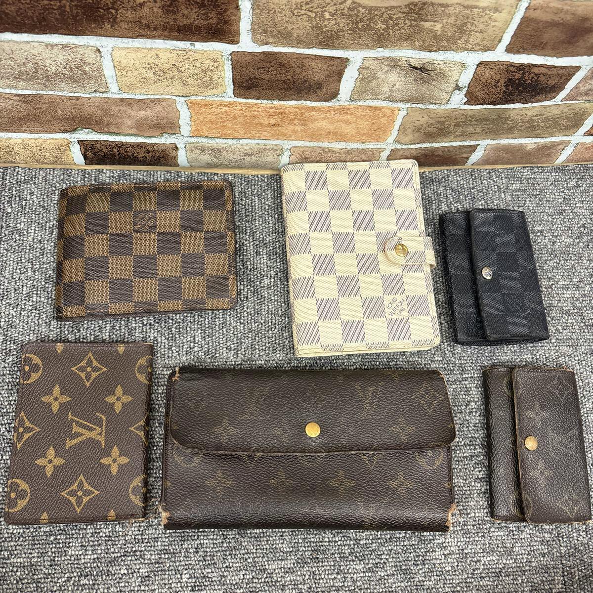Six Louis Vuitton small leather goods (wallets, key cases and card holders) arranged on a carpeted floor in front of a brick-patterned wall.