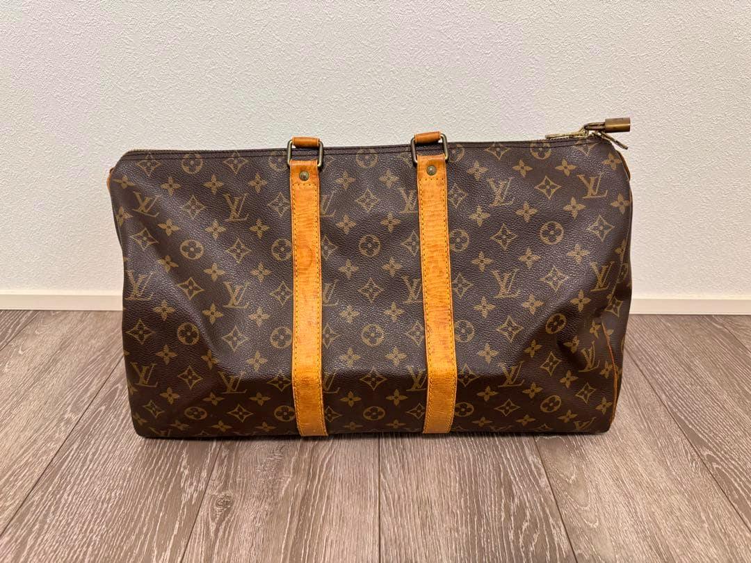 A Louis Vuitton monogram duffle bag in brown monogram canvas with cow leather straps placed on a wooden floor against a white wall.