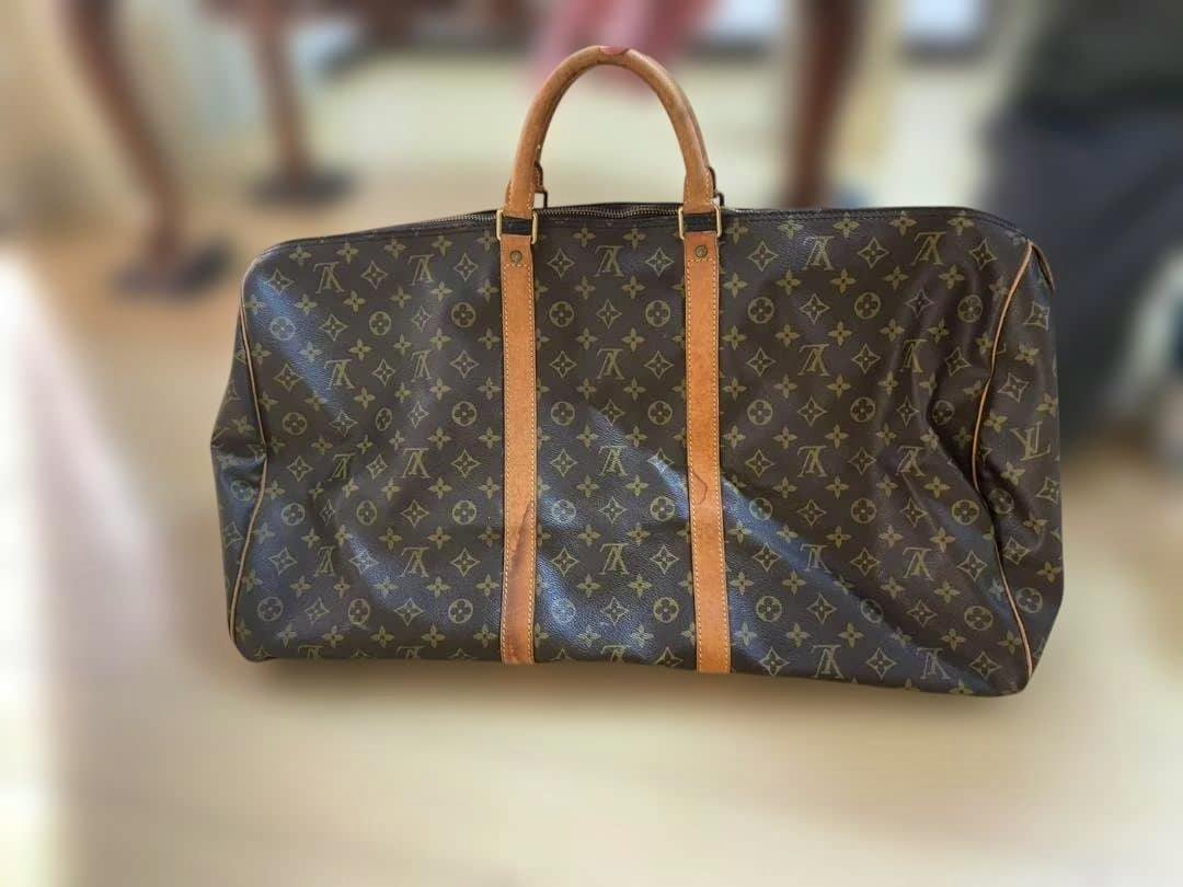 Front view of a brown Louis Vuitton monogram large duffle bag with tan leather handles and vertical leather straps on a neutral background.
