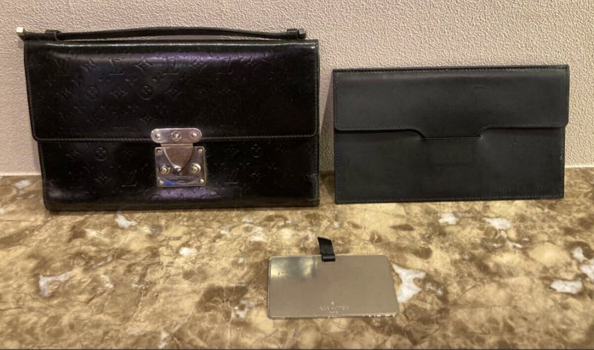 Front view of a black Louis Vuitton monogram clutch with metal clasp displayed next to a plain black clutch and a metal plate on a marble surface.
