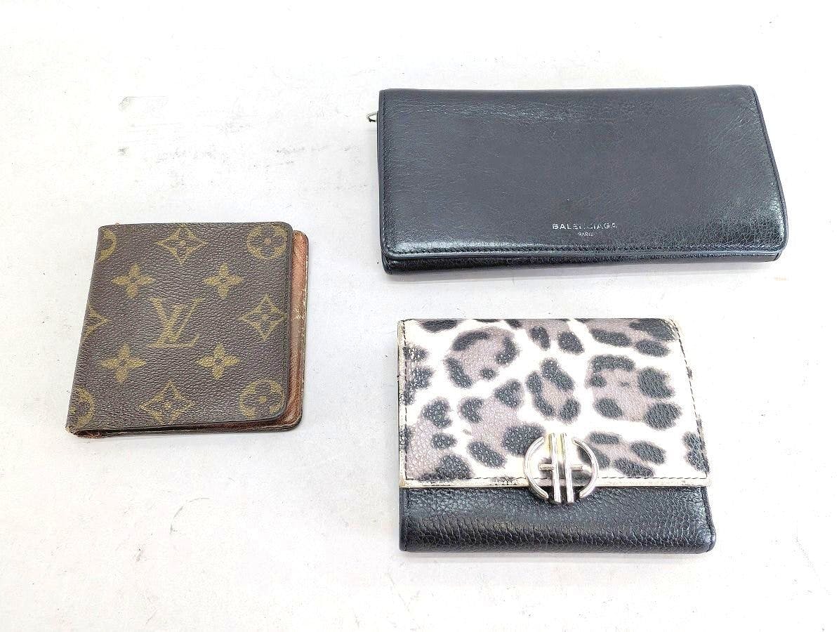 Three wallets on a white surface: a Louis Vuitton monogram wallet, a long black Balenciaga wallet, and a small leopard-print wallet.