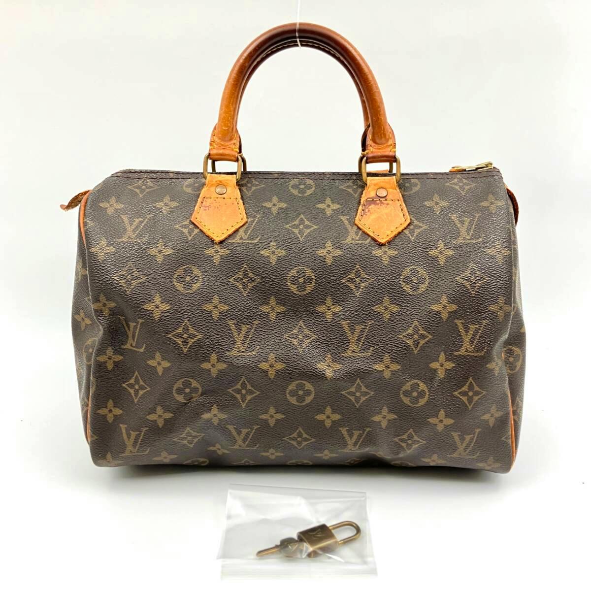 Front-facing LOUIS VUITTON monogram canvas Speedy bag with leather top handles and a small padlock and keys in a plastic bag placed in front.