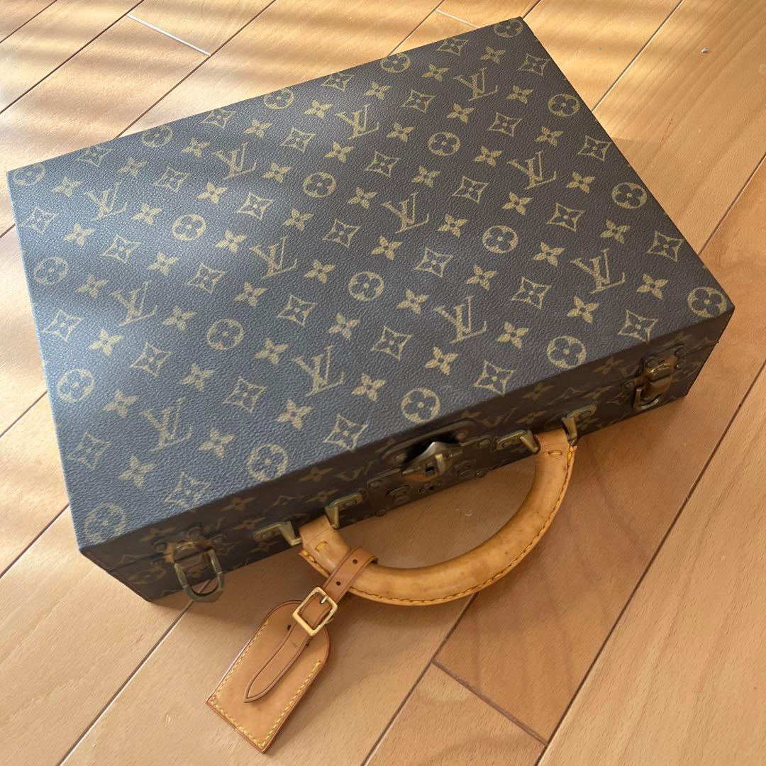 Closed Louis Vuitton monogram carry case with a handle and luggage tag resting on a wooden floor.