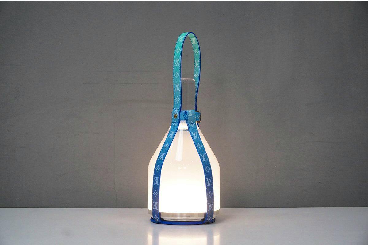 Blue Louis Vuitton Bell Lamp desk lamp with monogram straps lit on a table against a plain gray background.