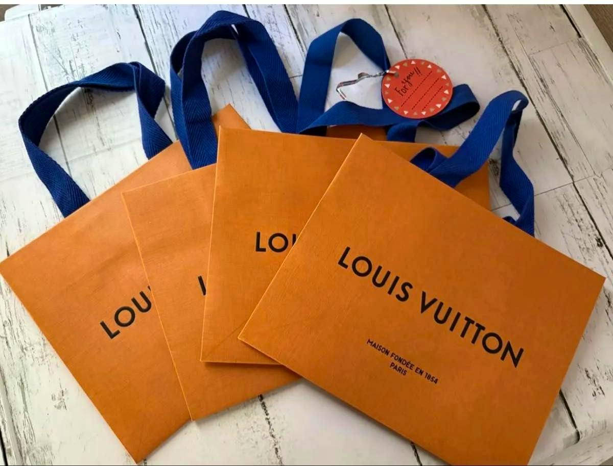 Four orange Louis Vuitton paper shopping bags with blue handles arranged on a white surface.