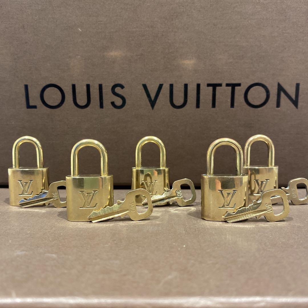 Five small gold Louis Vuitton padlocks with matching keys arranged on a brown surface in front of a Louis Vuitton box.
