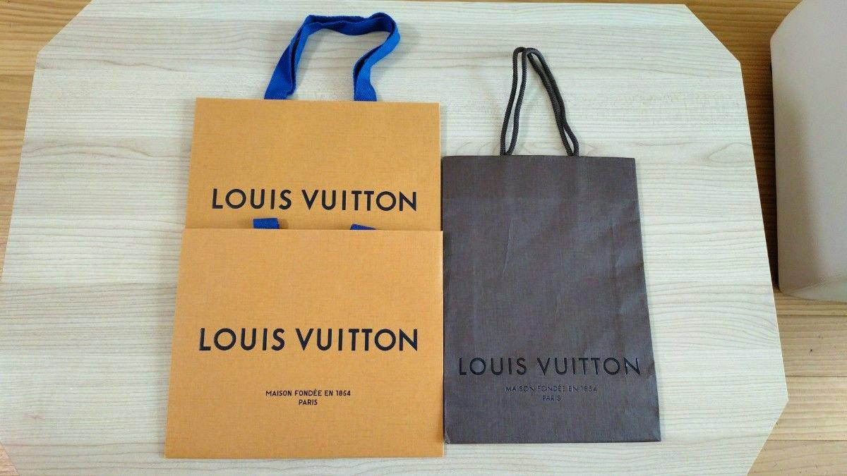 Three Louis Vuitton paper bags (two orange and one brown) laid flat on a light wood surface.