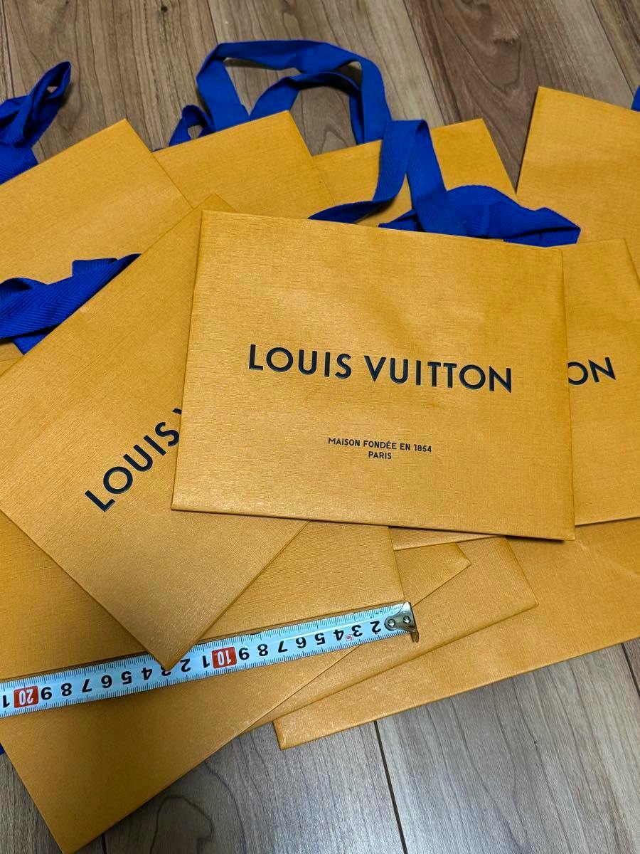 Orange Louis Vuitton paper shopping bags with blue ribbon handles arranged on a wooden floor with a measuring tape across them.