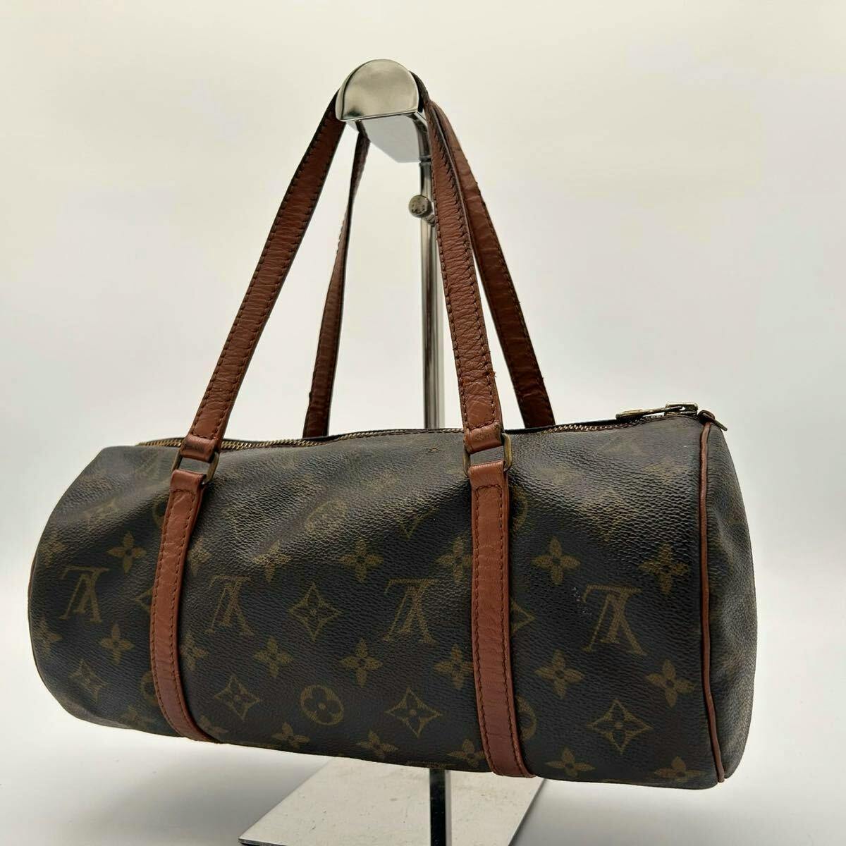 Louis Vuitton Papillon30 monogram cylindrical bag with brown leather handles displayed on a stand.