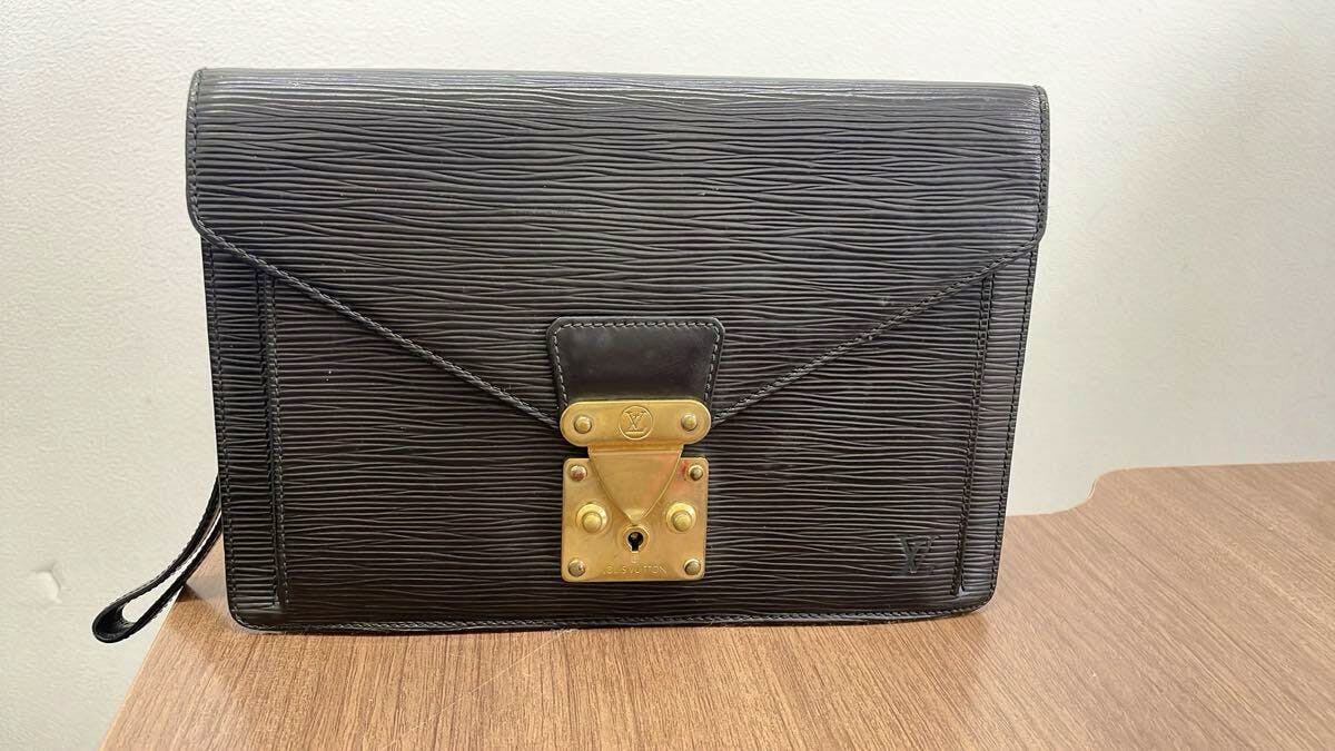 Front view of a black Louis Vuitton clutch bag in textured cow leather with a brass lock, shown on a wooden surface.