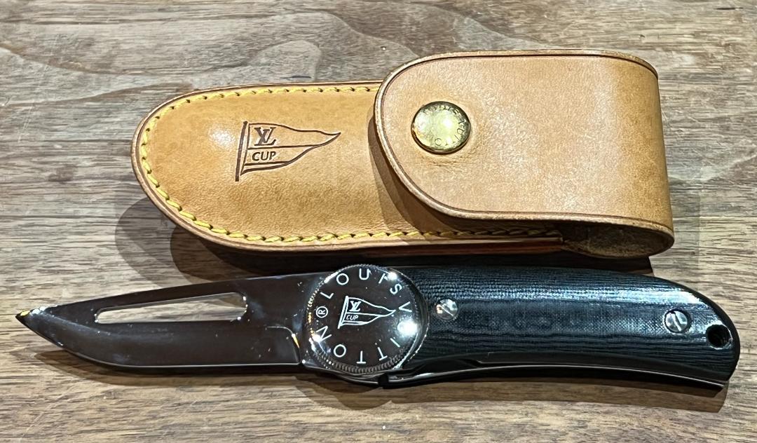 Louis Vuitton cup knife with its leather case placed on a wooden surface, the knife open showing the blade and black handle.