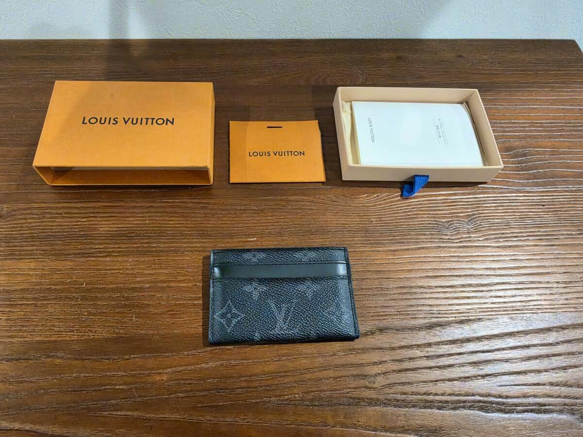 Louis Vuitton Monogram Eclipse card case displayed on a wooden table with its box and small packaging pieces.