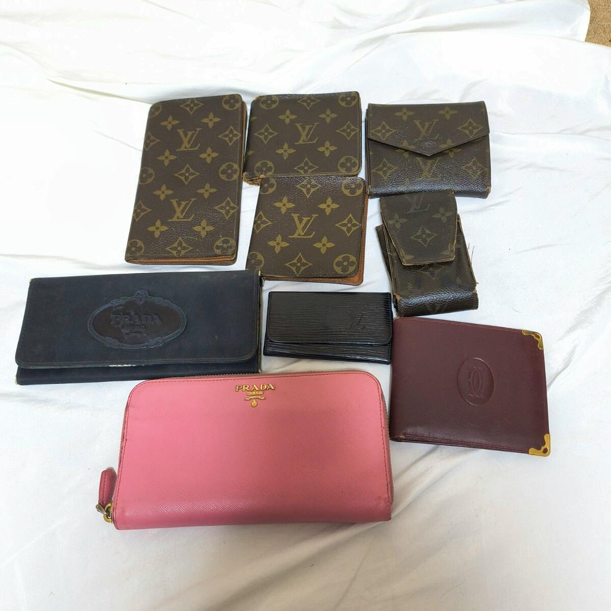 Nine branded wallets and small leather goods, including Louis Vuitton monogram pieces, a pink Prada zip wallet, and a burgundy Cartier wallet, arranged on a white cloth.