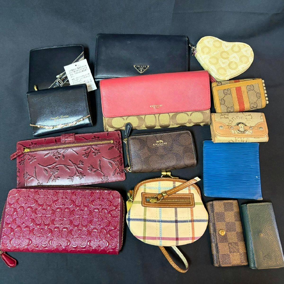 Multiple wallets, key cases, and small pouches (Louis Vuitton, Prada, Coach and others) arranged on a dark background.