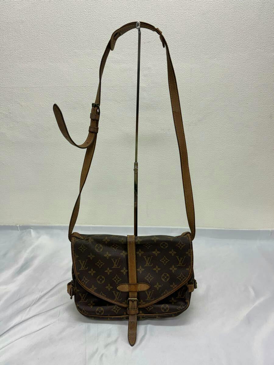 Front view of a Louis Vuitton monogram shoulder bag with flap and adjustable leather strap hanging from a stand.