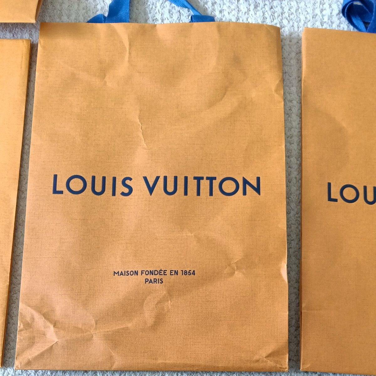 Orange Louis Vuitton paper shopper bag laid flat showing blue "LOUIS VUITTON" logo, smaller Paris text, and blue handles.