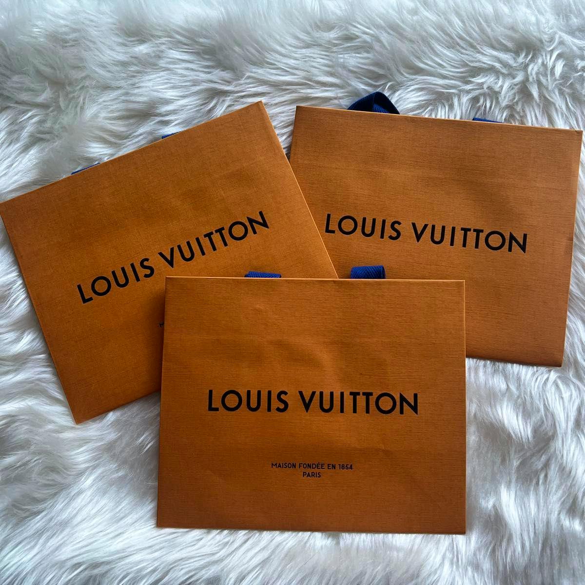 Three LOUIS VUITTON orange paper shopping bags with blue handles and black logo arranged on a white faux fur background.