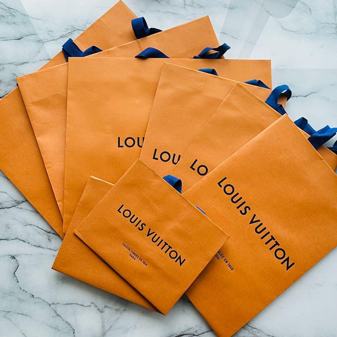 Multiple orange Louis Vuitton shopping bags with blue ribbon handles arranged on a marble surface.