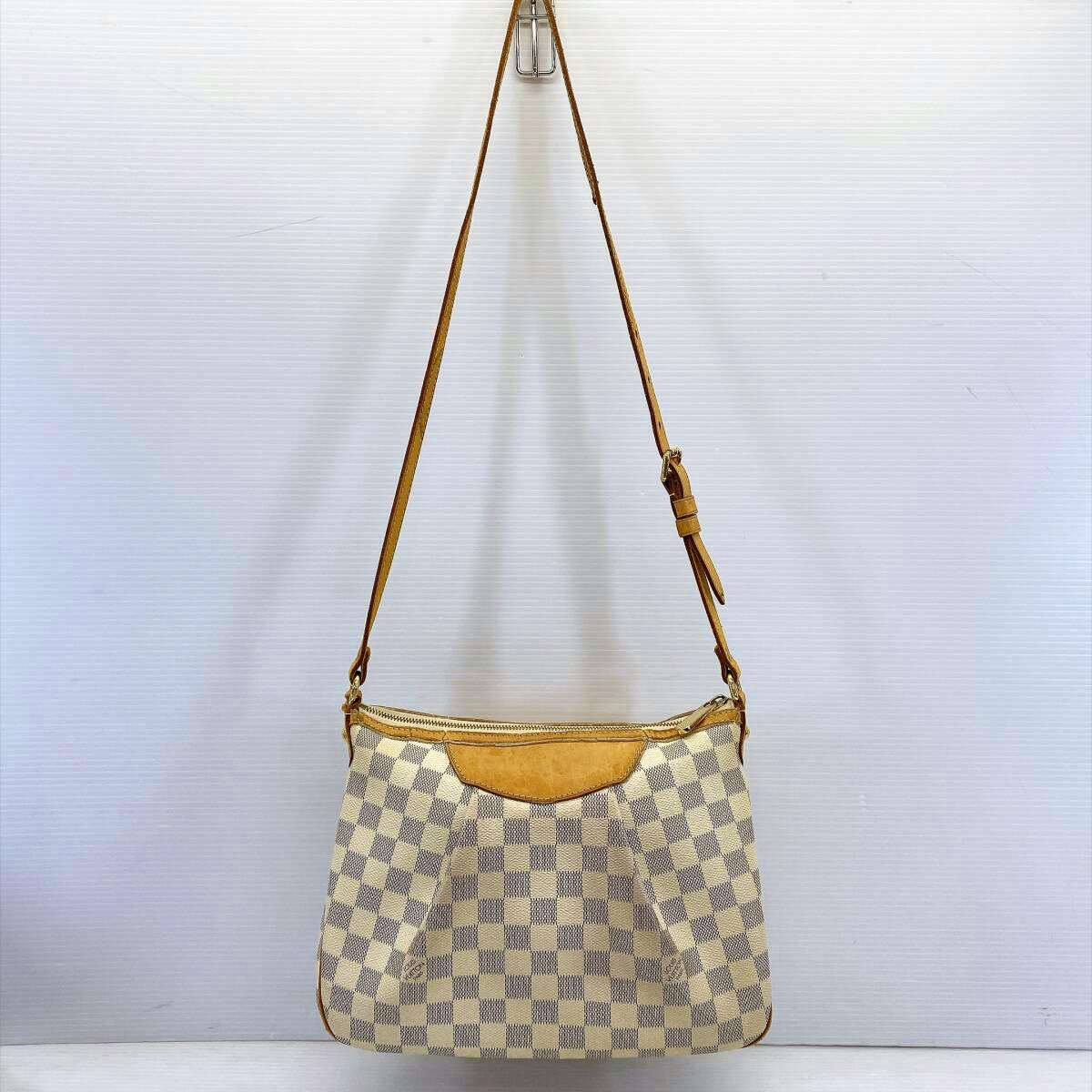 Front view of the Louis Vuitton Damier Azur Siracusa PM canvas shoulder bag with vachetta leather trim and adjustable strap hanging against a white background.