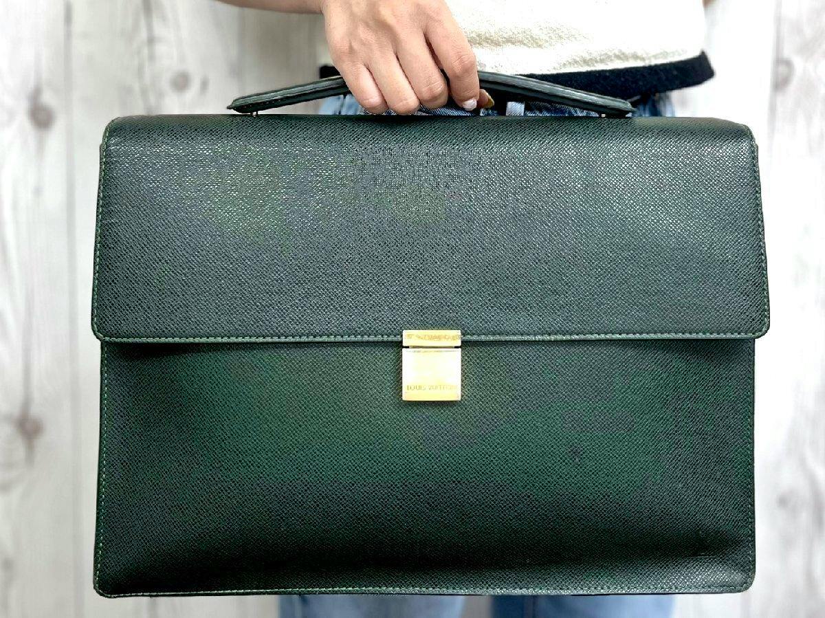 Front view of a dark green Louis Vuitton leather business bag with a top handle and gold-tone clasp being held by a person.