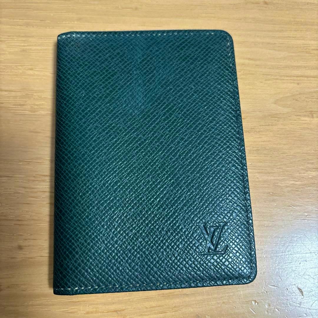 Green Taiga cow leather card case on a wooden surface showing the front with an embossed LV logo.
