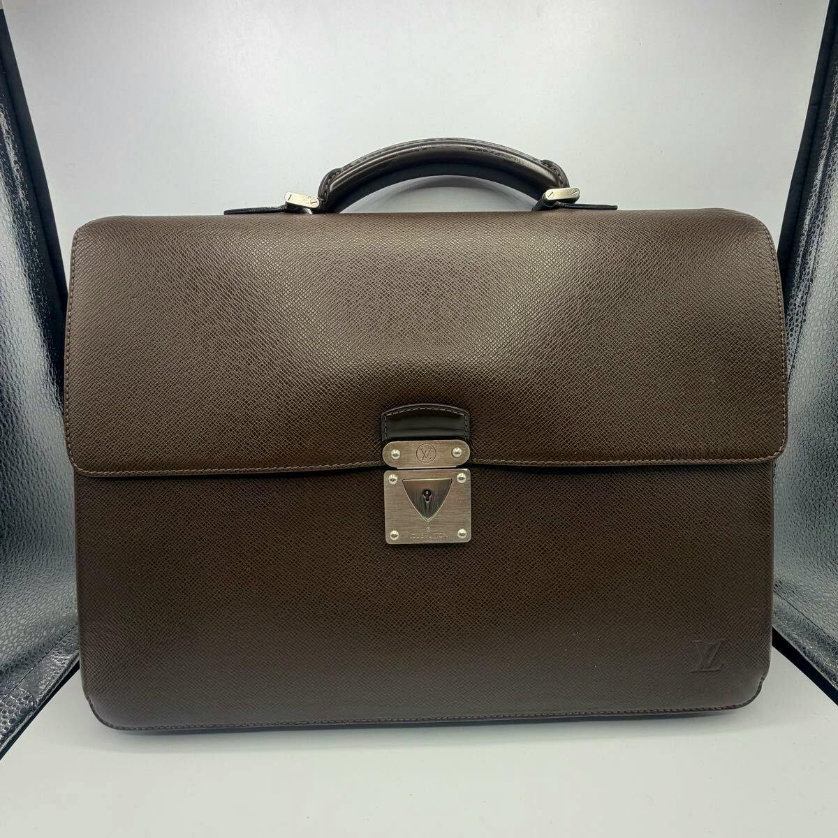 Front view of a brown Louis Vuitton Taiga leather briefcase with a top handle and central metal lock.