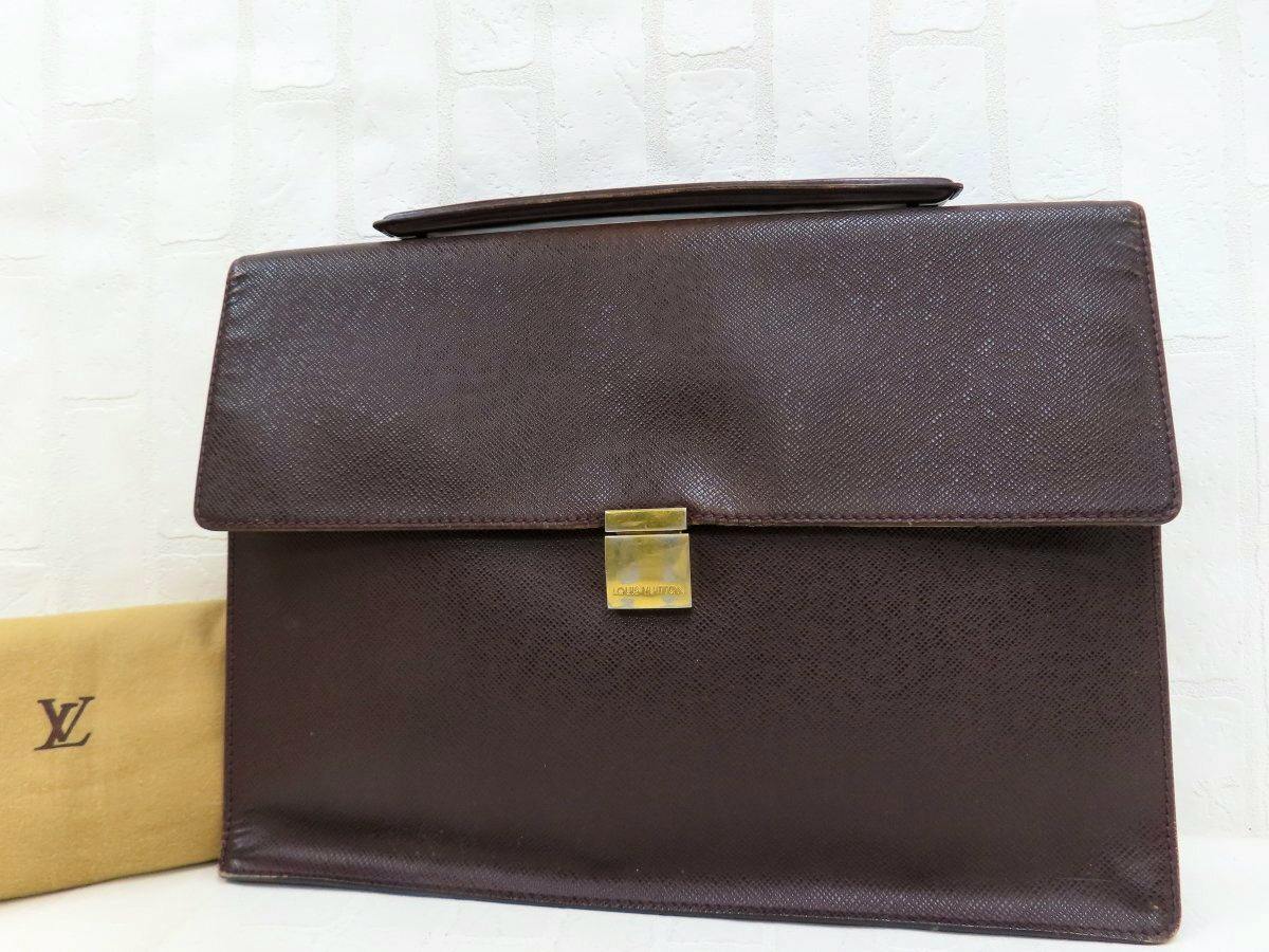 Front view of a brown Louis Vuitton cow leather business bag with top handle and gold clasp, with part of a dust bag visible.