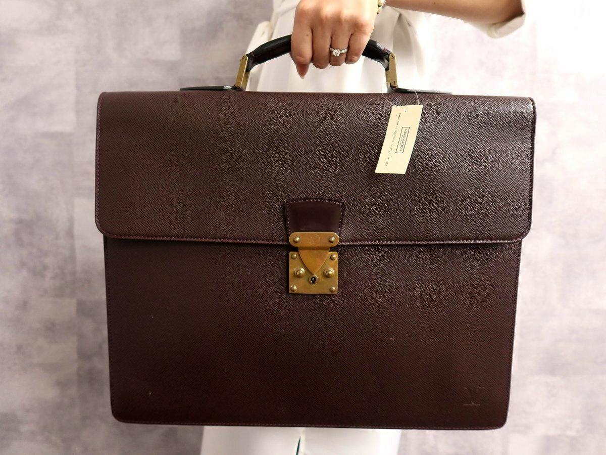 Front view of the Louis Vuitton brown leather men's briefcase (Taiga Serviette Moscova) being held by a hand.