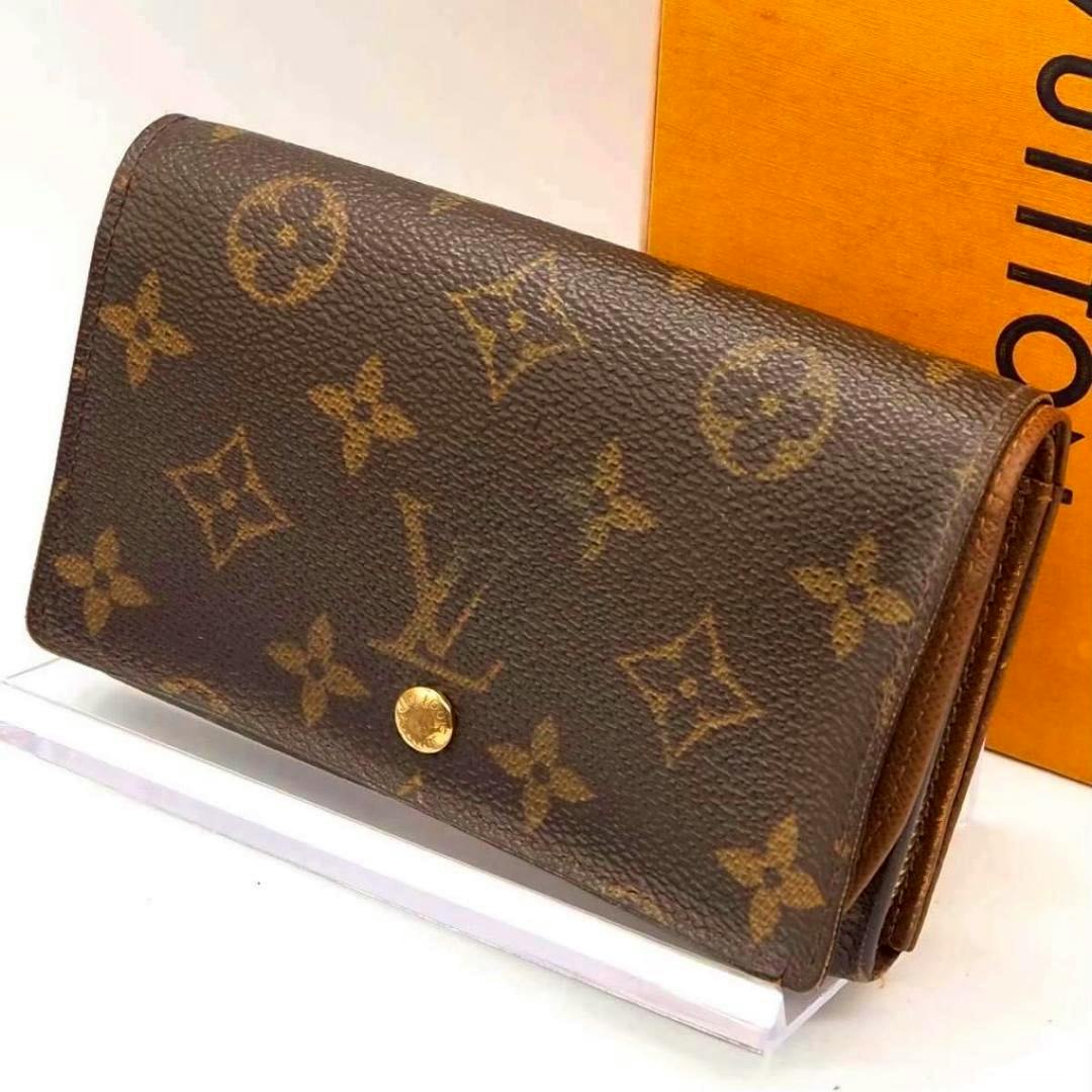 A Louis Vuitton Tresor Monogram canvas compact bi-fold wallet shown folded from the front with its gold snap button and part of an orange box visible behind it.