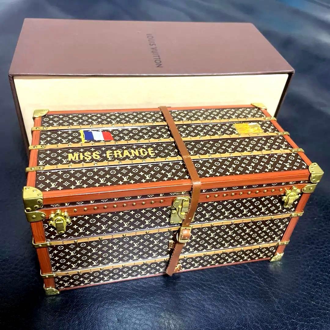 Trunk-shaped Louis Vuitton monogram paperweight with metal corner fittings shown in front of its box.
