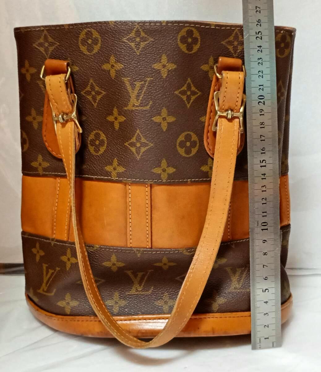 Front view of the Louis Vuitton monogram canvas bucket bag with tan leather trim, shoulder straps, and a ruler for scale.