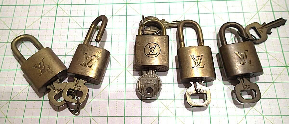 Six Louis Vuitton padlocks with keys arranged on a green 1 cm grid mat.