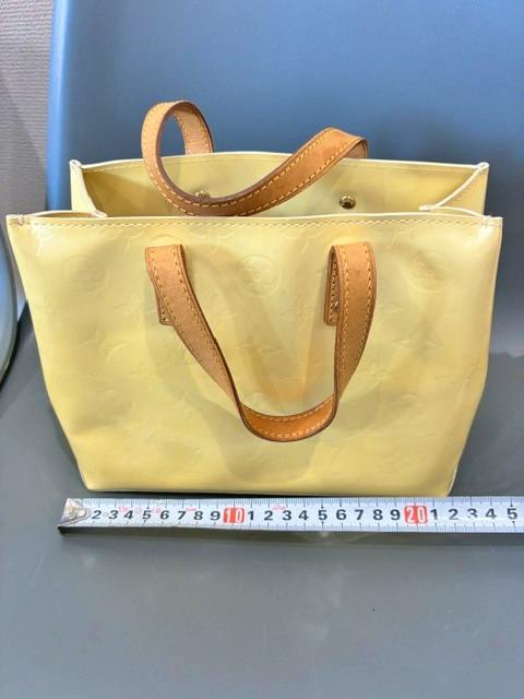 Front view of a yellow Louis Vuitton Vernis handbag with brown leather handles shown next to a measuring tape.