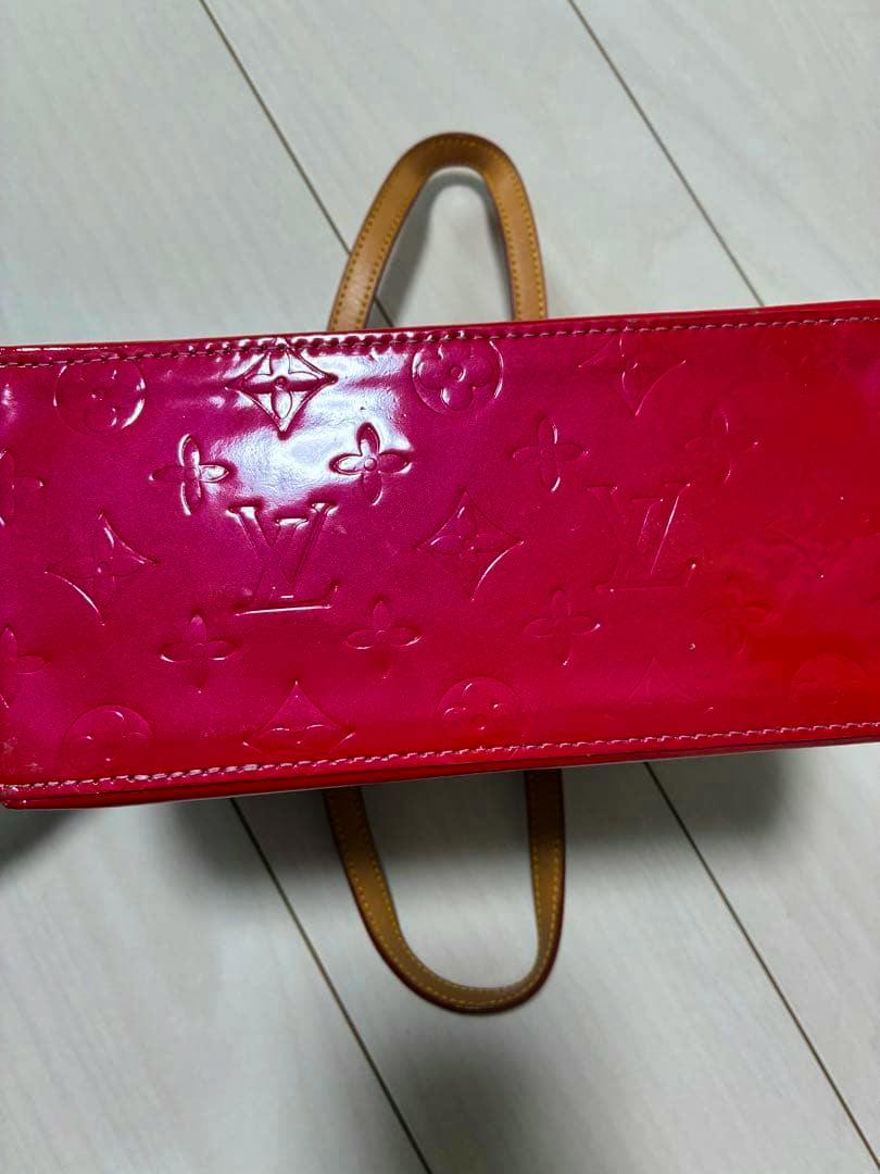 Red Louis Vuitton Vernis handbag on a light wood floor showing embossed monogram pattern and tan leather handles.