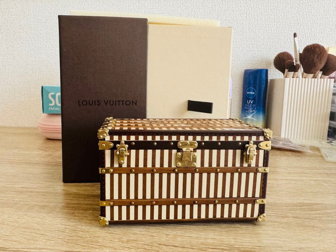 Wooden Louis Vuitton trunk ornament with striped panels and brass hardware placed on a table with its box behind.