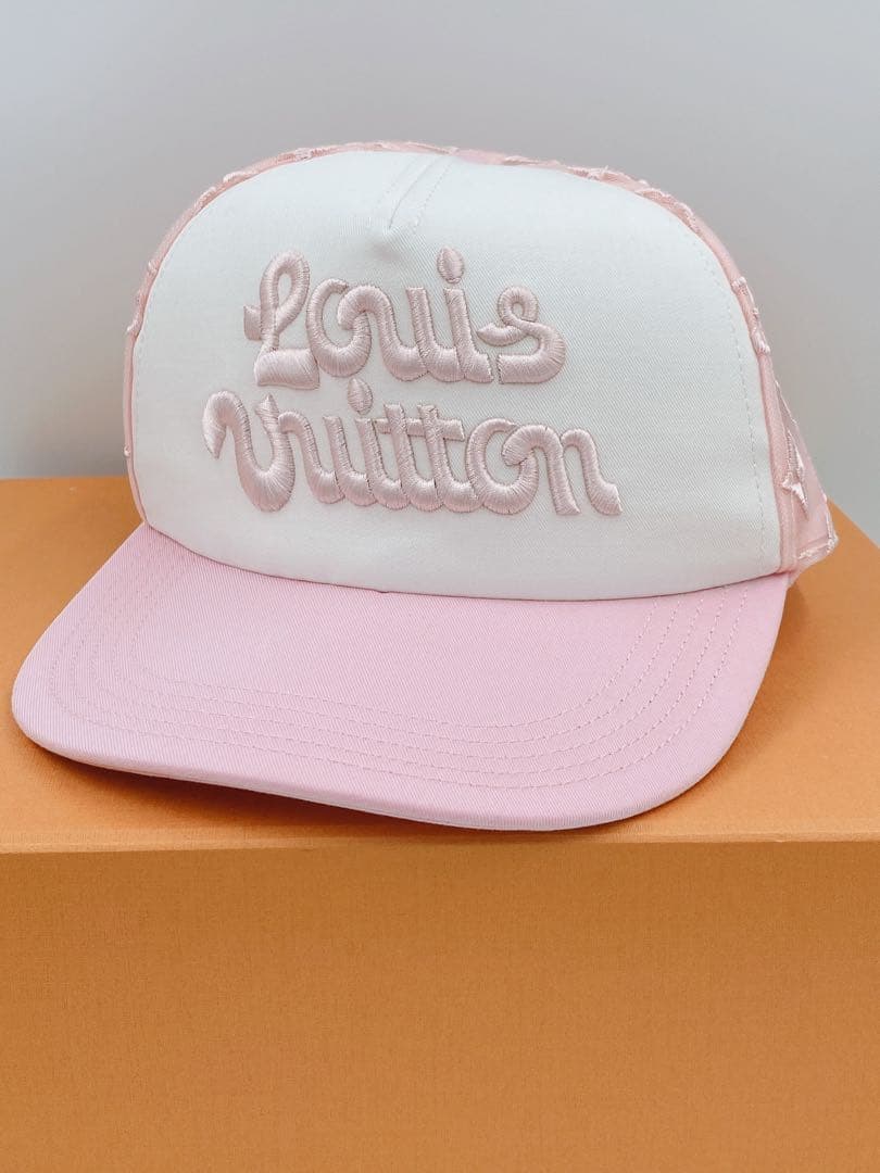 Front view of a pink and white Louis Vuitton hat with "Louis Vuitton" embroidered on the front, placed on an orange box.