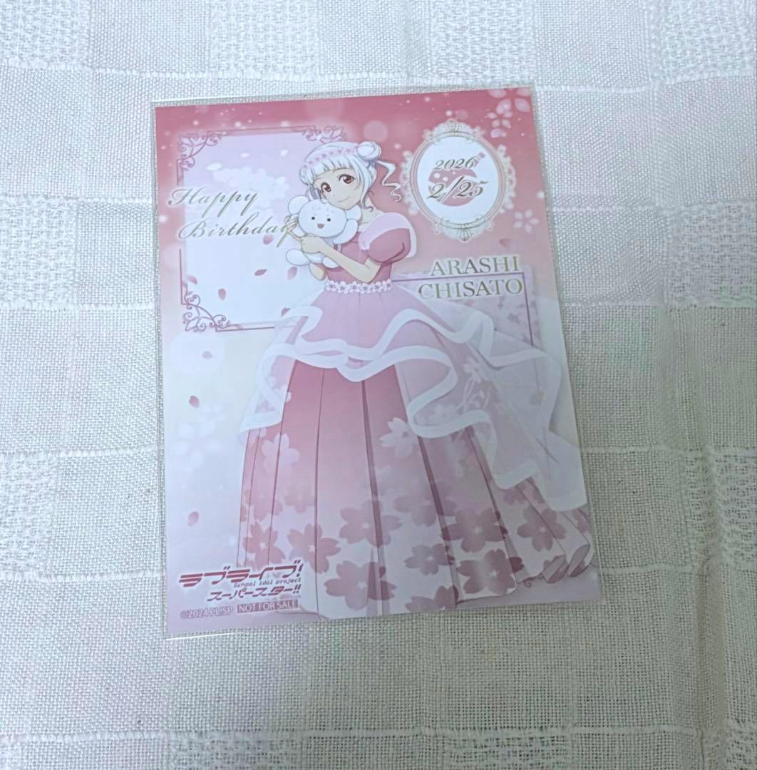 Photocard of Arashi Chisato in a pink dress holding a stuffed animal with "Happy Birthday" text and a date, placed on a fabric surface.