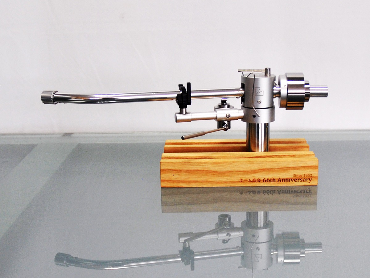 A silver record player tone arm mounted on a wooden block, shown in profile with its reflection on glass.