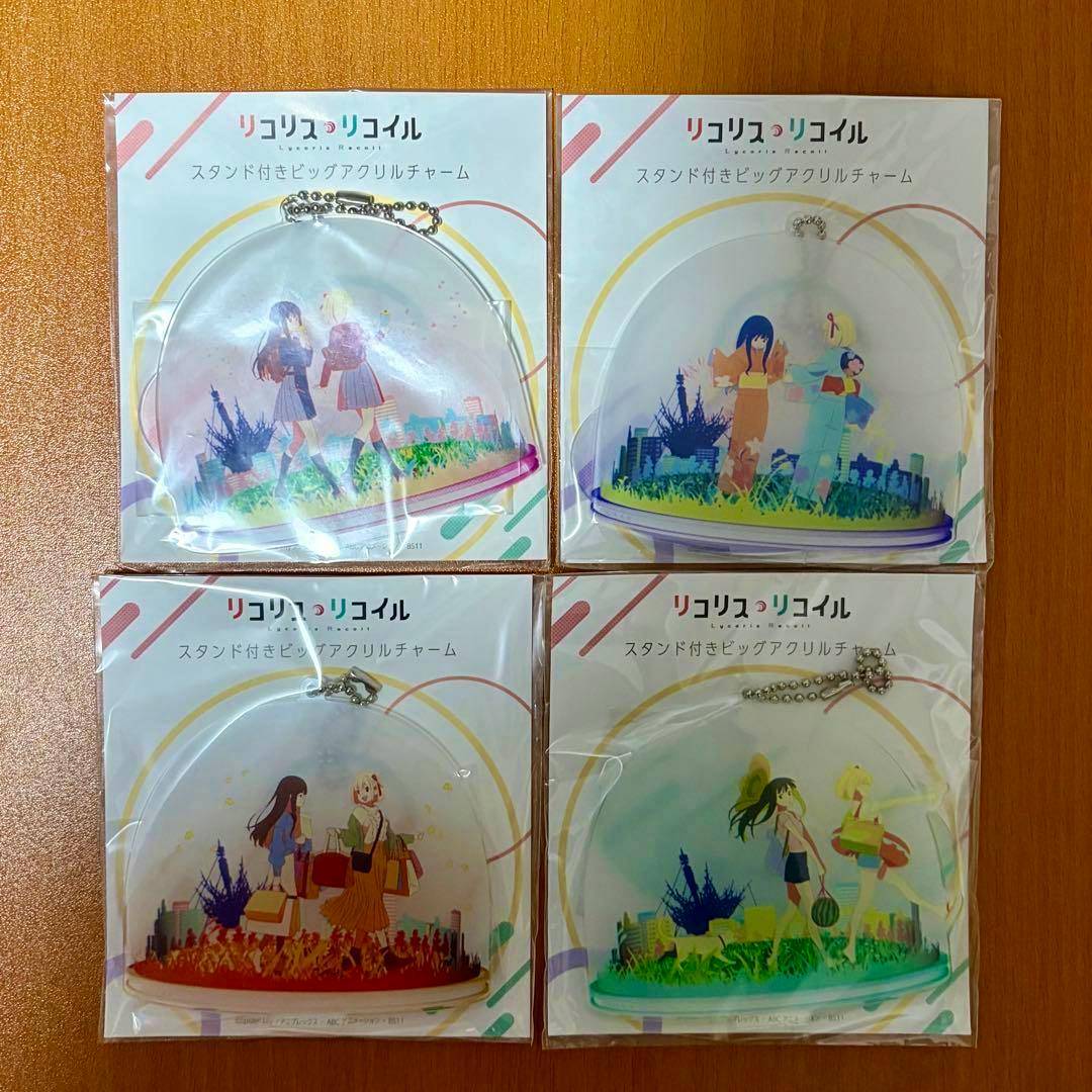 Front views of four packaged Lycoris Recoil stand-attached big acrylic charms (keychains) arranged in a 2x2 grid on a wooden background.