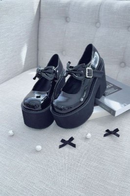 Black synthetic leather platform pumps with large ribbon bows placed on a light gray tufted couch.