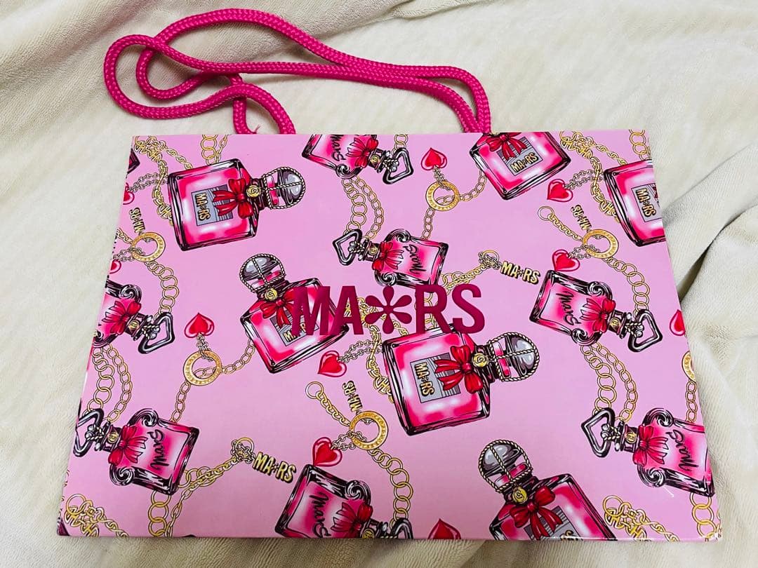 Front of a pink MA*RS paper shopper bag showing perfume-bottle and chain pattern with pink rope handles.