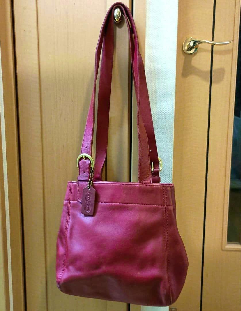 Red OLD COACH cow-leather shoulder bag with straps and leather tag hanging on a door.