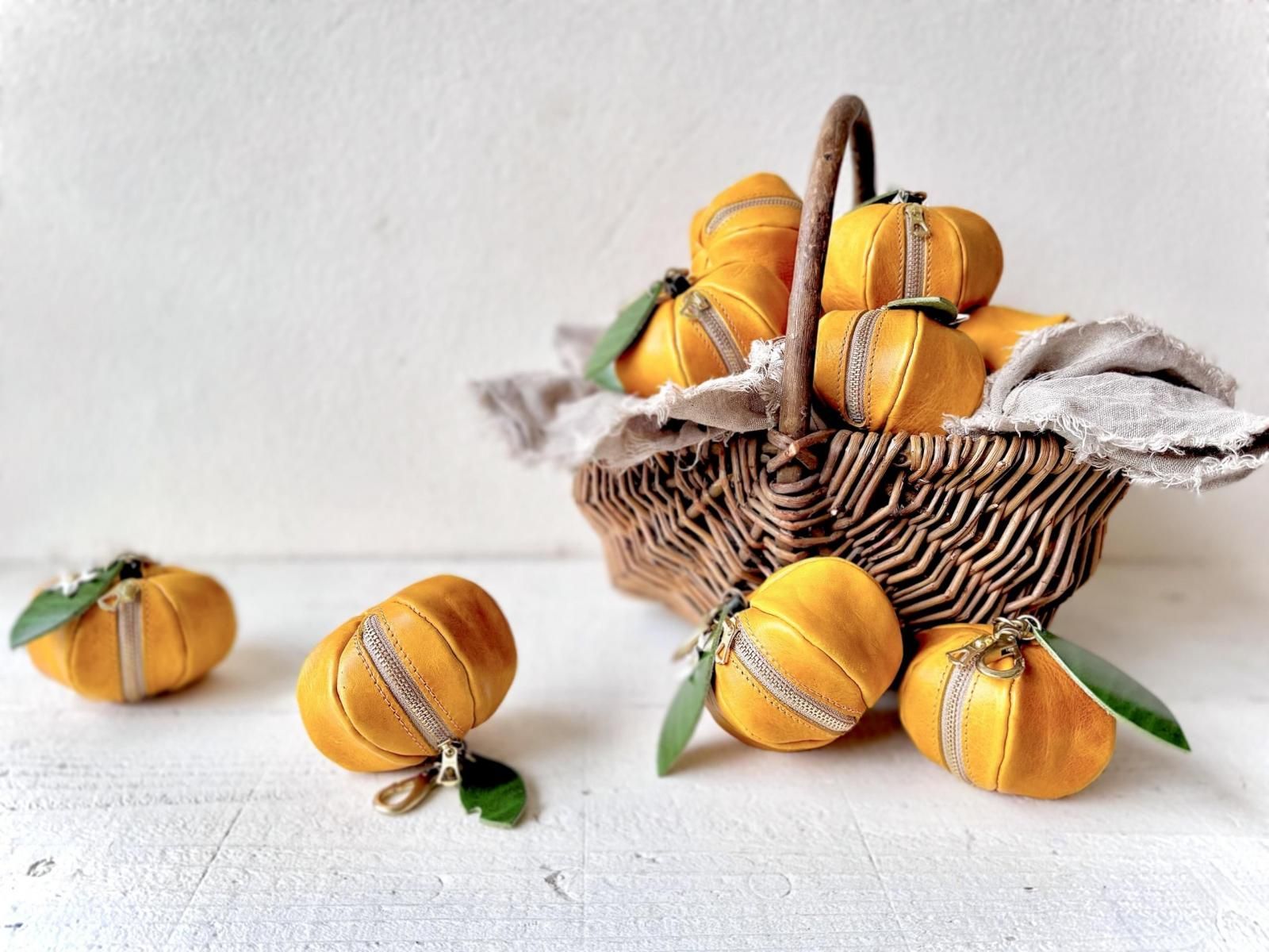 Yellow-orange Italian leather mini pouches shaped like mandarins with zippers and green leaf accents arranged around a wicker basket.
