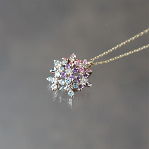 Close-up of the hydrangea pendant head with multiple small colored gemstones on a short chain against a neutral reflective background.