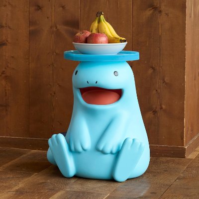 Blue plastic mini table shaped like the 'ヌオー' character with a flat top holding a bowl of fruit on a wooden floor.