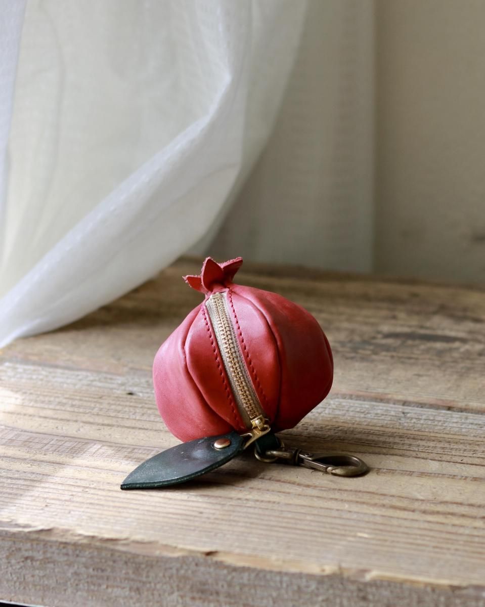 Red cow-leather mini pouch shaped like a pomegranate with a green leather leaf zipper pull and brass clasp sitting on a wooden surface.