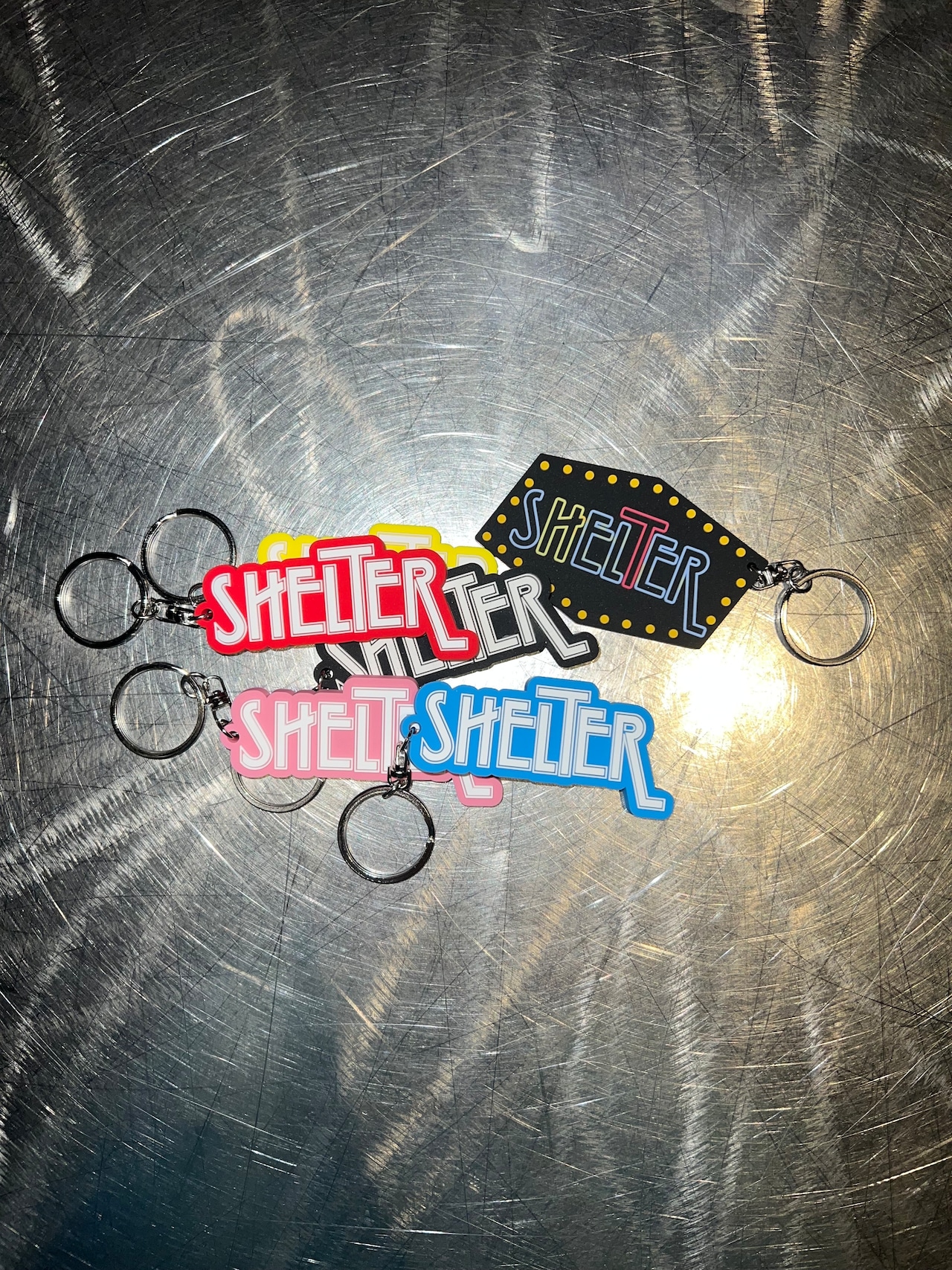 Four rubber keychains reading "SHELTER" in different colors laid on a brushed metal surface.