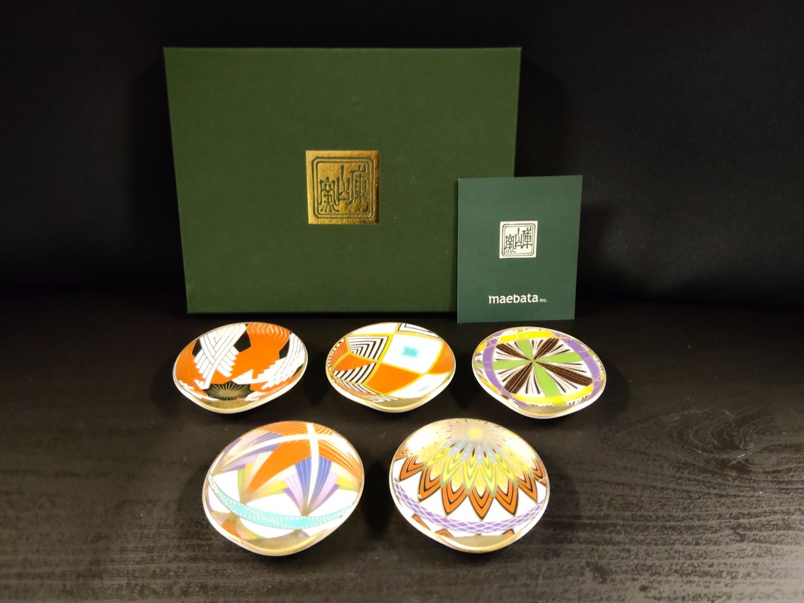 Five small decorative ceramic plates with colorful temari patterns arranged in front of a green presentation box.
